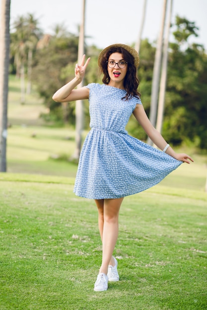 La muchacha linda divertida con un vestido azul cielo está de pie en un parque tropical. La chica parece sorprendida.