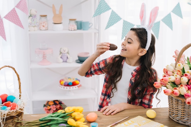 Muchacha linda sonriente con los oídos del conejito sobre su cabeza que come el huevo de Pascua del chocolate