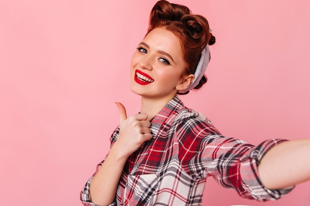 Foto gratuita muchacha modela atractiva que muestra el pulgar para arriba. disparo de estudio de jengibre joven haciendo selfie en espacio rosa.