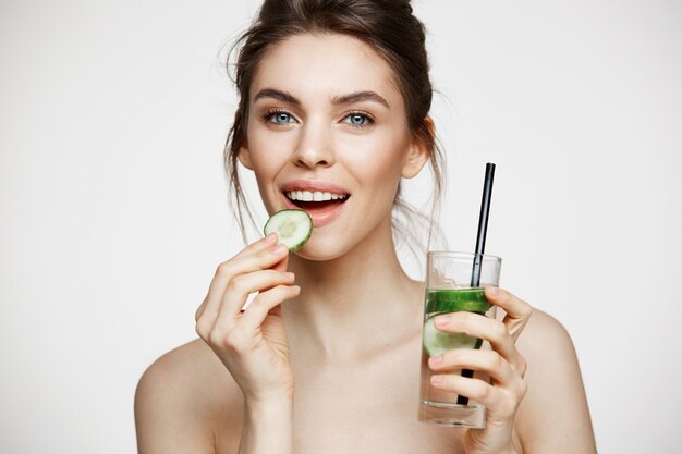 Muchacha morena joven positiva que sonríe mirando la cámara que come la rebanada del pepino que sostiene el vidrio de agua sobre el fondo blanco. Belleza y salud.