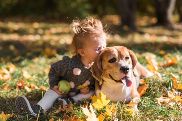 Muchacha del niño que besa a su perro que se sienta en hierba en el bosque