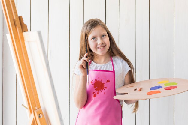 Muchacha pensativa sonriente que sostiene la paleta y el cepillo de pintura que se colocan cerca del caballete