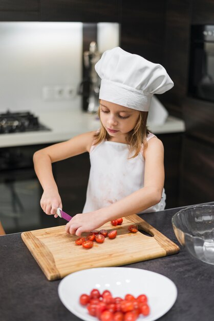 Muchacha pequeña que lleva el sombrero del cocinero y los tomates del corte del delantal en tajadera en cocina