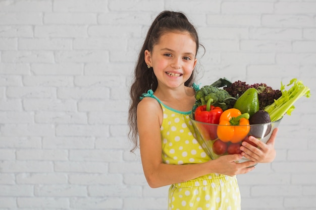 Muchacha que está parada delante de la pared blanca que sostiene el cuenco vegetal