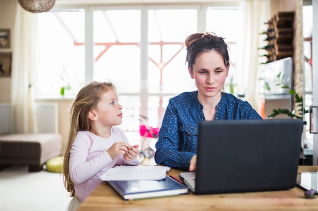 Muchacha que mira a su madre que trabaja en la computadora portátil sobre el escritorio de madera