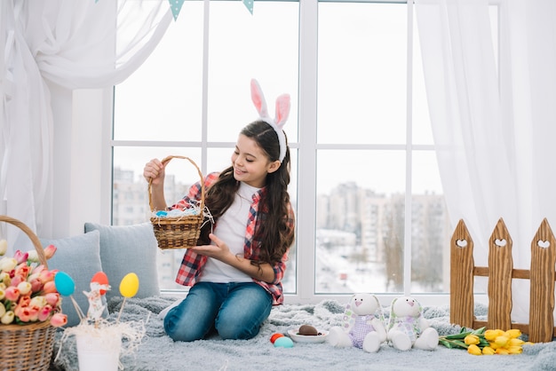 Muchacha que se sienta en la cama que mira la cesta de los huevos de Pascua en casa