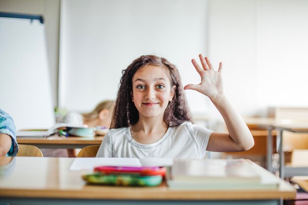 Muchacha que se sienta en el escritorio de la escuela que agita la mano