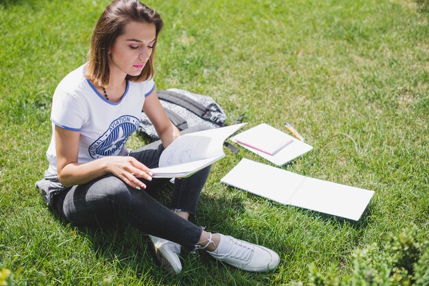 Muchacha que se sienta en el parque que sostiene la lectura del cuaderno