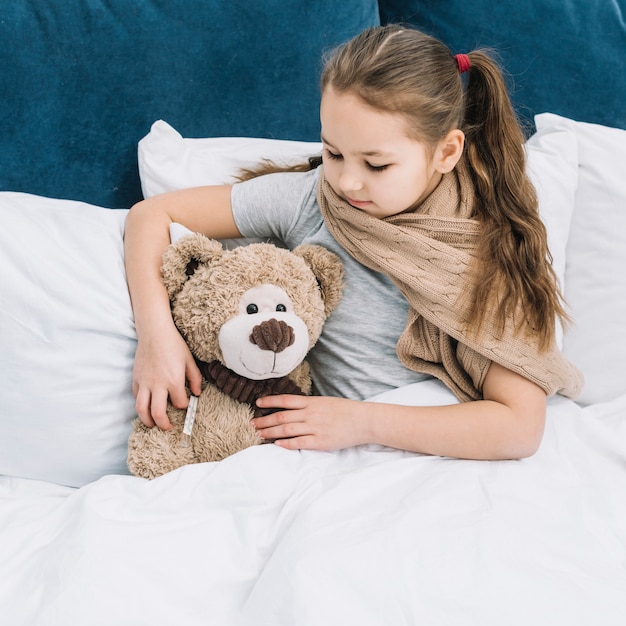 Foto gratuita muchacha que tiene fiebre que sostiene el termómetro en la mano del oso de peluche que controla la temperatura