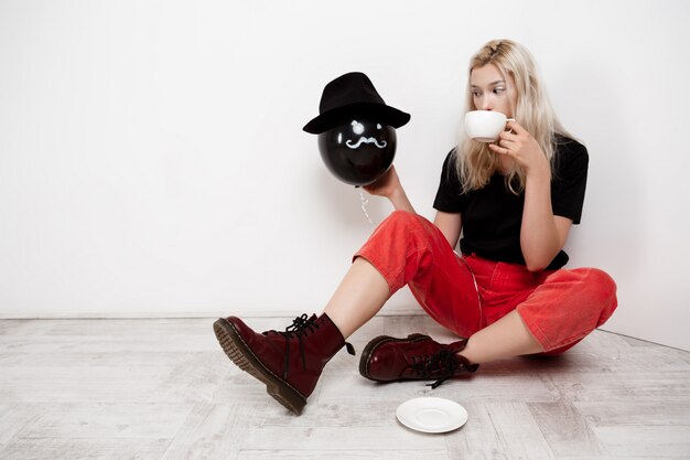 Muchacha rubia hermosa joven que sostiene el globo negro en el sombrero que se sienta en el piso que bebe el café sobre la pared blanca.