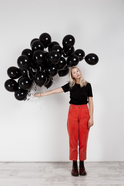 Muchacha rubia hermosa joven que sostiene globos negros sobre la pared blanca.