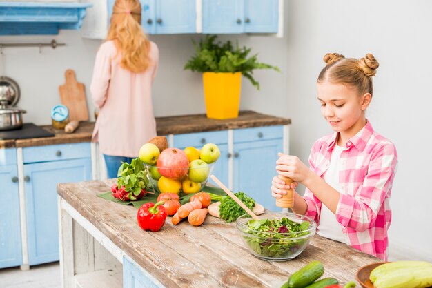 Muchacha rubia que muele la pimienta en cuenco de ensalada con su madre en la cocina