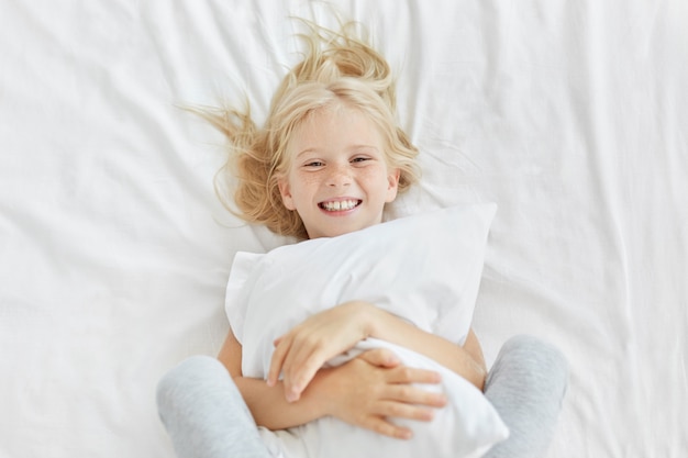 Muchacha rubia sonriente que abraza la almohada blanca mientras que está en la guardería, tiene buen humor mientras ve a alguien y se acuesta en la cama blanca. Pequeña niña adorable que tiene hora de acostarse. Concepto de descanso