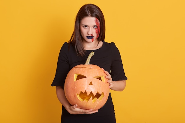 Muchacha seria con la expresión facial enojada que se coloca con la calabaza en manos en el estudio aislado en amarillo, hembra atractiva con la herida sangrienta en su cara, concepto de Halloween.