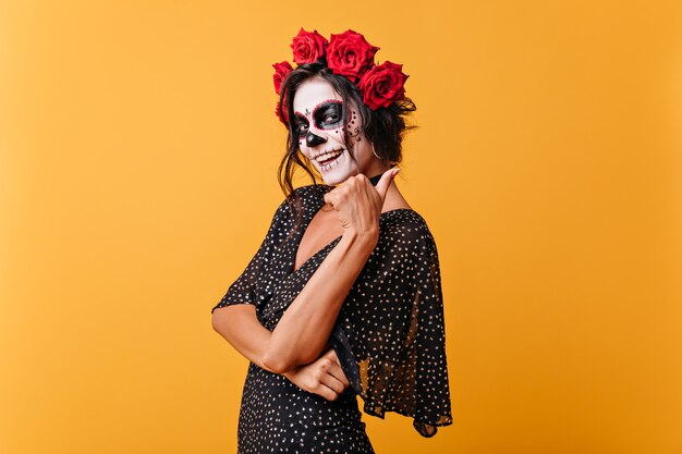 La muchacha sonriente con el arte de la cara para el carnaval muestra el pulgar hacia arriba, posando en traje negro sobre fondo aislado.