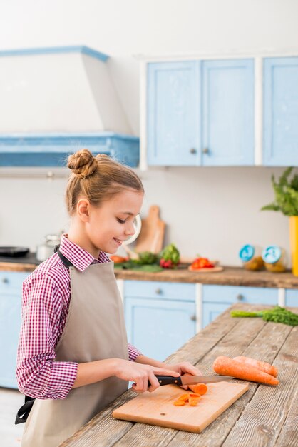 Muchacha sonriente que corta la zanahoria con el cuchillo en tajadera en la cocina