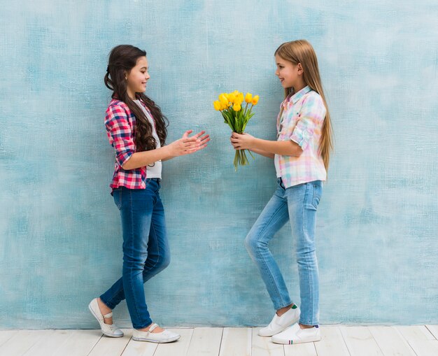 Muchacha sonriente que da la flor amarilla del tulipán a su amigo contra la pared azul