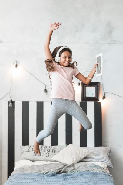 Muchacha sonriente que lleva el auricular que salta sobre la cama con la tableta digital