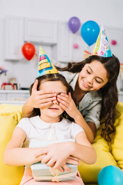 Muchacha sonriente que oculta los ojos de su amigo que sostienen la caja de regalo en su mano