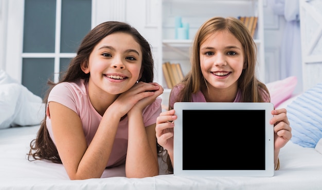 Muchacha sonriente que pone con su amigo en la cama que muestra la tableta digital de la pantalla en blanco