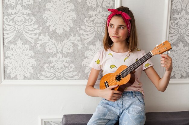 Muchacha sonriente que se sienta en el sofá que ajusta el ukelele y el pensamiento