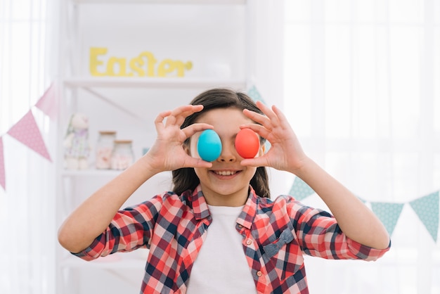 Muchacha sonriente que sostiene los huevos de Pascua rojos y azules sobre sus ojos en casa