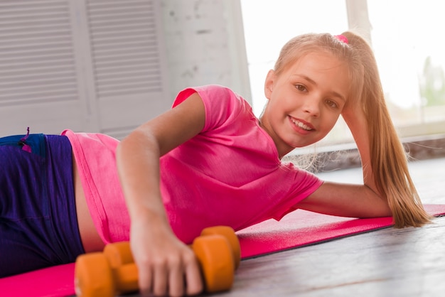 Muchacha sonriente rubia que miente en la estera rosada del ejercicio con una pesa de gimnasia anaranjada