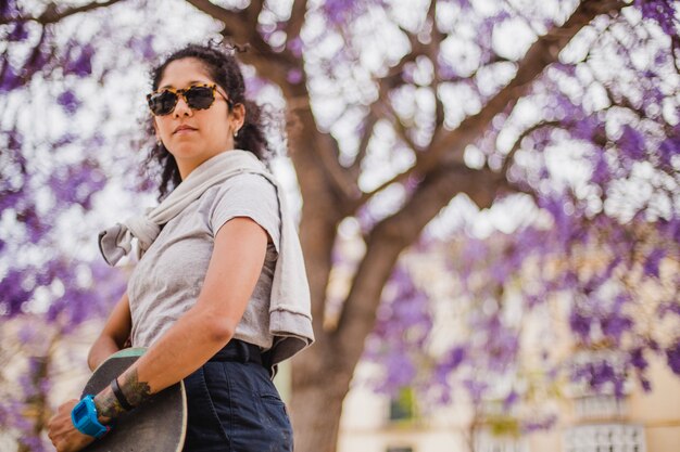 Muchacha triguena en gafas de sol que sostienen el monopatín de pie