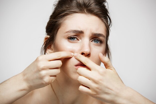 La muchacha triguena joven disgustó de su piel de la cara del acné del problema sobre el fondo blanco. Salud cosmetología y cuidado de la piel.