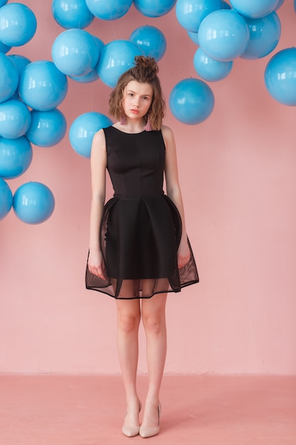 Muchacha triste en vestido negro lindo en fondo rosado con los globos azules