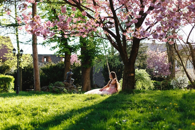 La muchacha en vestido rosado se sienta debajo del árbol floreciente de Sakura en el parque