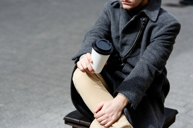 El muchacho en abrigo gris sostiene la taza de café en su rodilla que se sienta en la calle