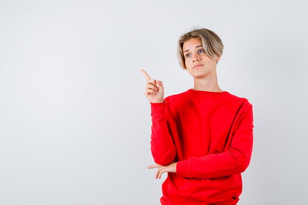 Muchacho adolescente apuntando hacia arriba con suéter rojo y mirando preocupado. vista frontal.