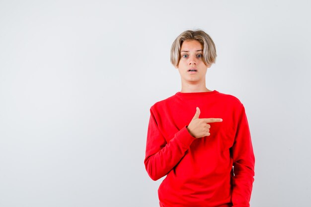 Muchacho adolescente apuntando hacia el lado derecho, abriendo la boca con un suéter rojo y mirando sorprendido, vista frontal.