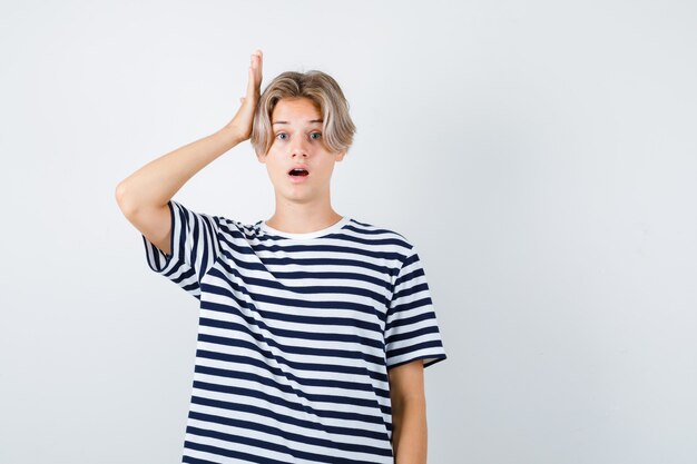 Muchacho adolescente en camiseta manteniendo la mano en la cabeza mientras abre la boca y mira sorprendido, vista frontal.
