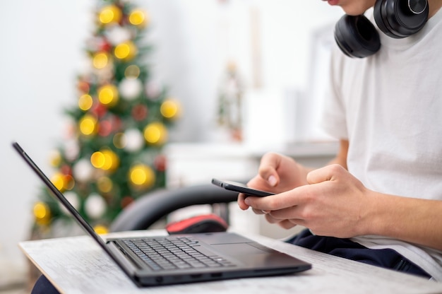 Muchacho adolescente está utilizando un teléfono inteligente con auriculares, portátil de rodillas en casa. Árbol de navidad en la pared