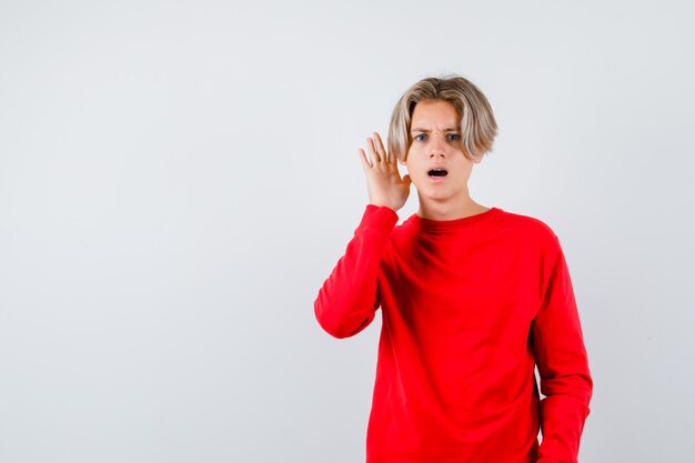 Muchacho adolescente joven con la mano cerca de la oreja en suéter rojo y mirando confundido. vista frontal.