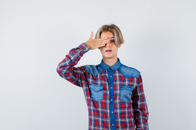 Muchacho adolescente joven con la mano en el ojo en camisa a cuadros y mirando se preguntó. vista frontal.