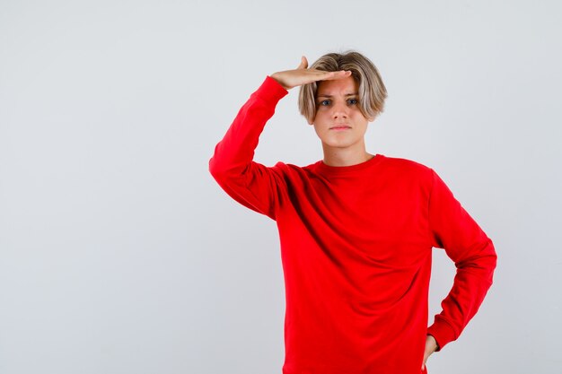 Muchacho adolescente joven con las manos sobre la cabeza en suéter rojo y mirando confundido. vista frontal.