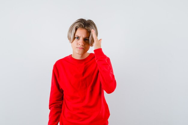 Muchacho adolescente joven con las manos sobre la cabeza para ver claramente en suéter rojo y mirando confundido, vista frontal.