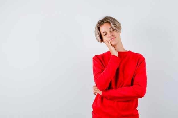 Muchacho adolescente joven que se inclina la mejilla en la mano en un suéter rojo y que parece relajado, vista frontal.