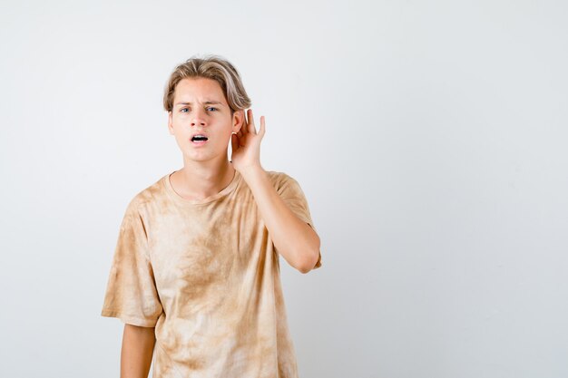 Muchacho adolescente joven que mantiene la mano detrás de la oreja en la camiseta y que mira perplejo, vista frontal.