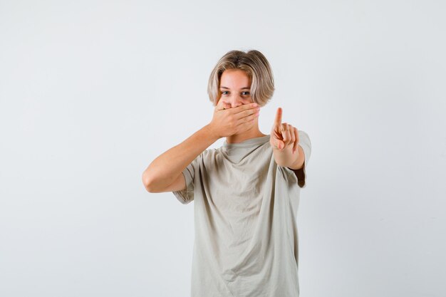 Muchacho adolescente joven que muestra espera en un gesto de minuto, manteniendo la mano en la boca en la camiseta. vista frontal.