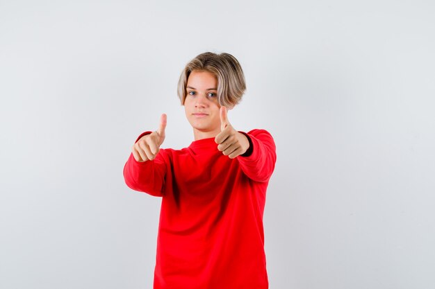 Muchacho adolescente joven que muestra los pulgares dobles para arriba en el suéter rojo y que parece complacido, vista frontal.