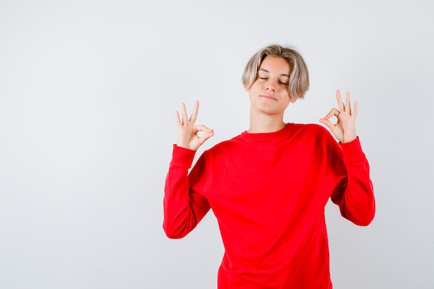 Muchacho adolescente joven que muestra el signo de mudra, manteniendo los ojos cerrados en un suéter rojo y luciendo relajado. vista frontal.