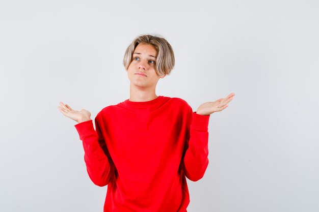 Muchacho adolescente joven en suéter rojo que muestra un gesto de impotencia y que mira pensativa, vista frontal.