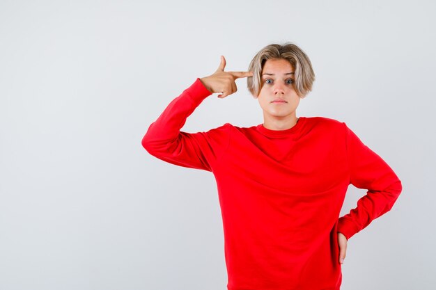 Muchacho adolescente joven en suéter rojo que muestra el gesto del suicidio y que mira perplejo, vista frontal.