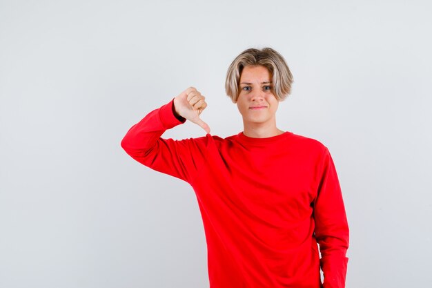 Muchacho adolescente joven en suéter rojo que muestra el pulgar hacia abajo y que mira disgustado, vista frontal.