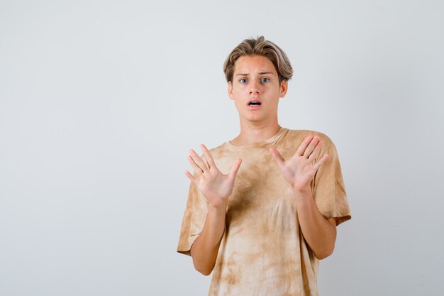 Muchacho adolescente levantando las manos para defenderse en camiseta y mirando aterrorizado, vista frontal.