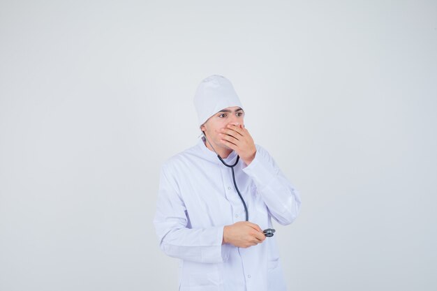 muchacho adolescente con la mano en la boca en uniforme de médico y mirando desconcertado